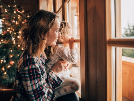 Woman holding young child up to look out window of home with Christmas tree in background