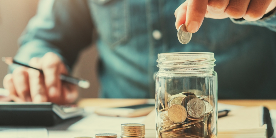 Man Holding Coin Above Jar While Using A Calculator In The Background